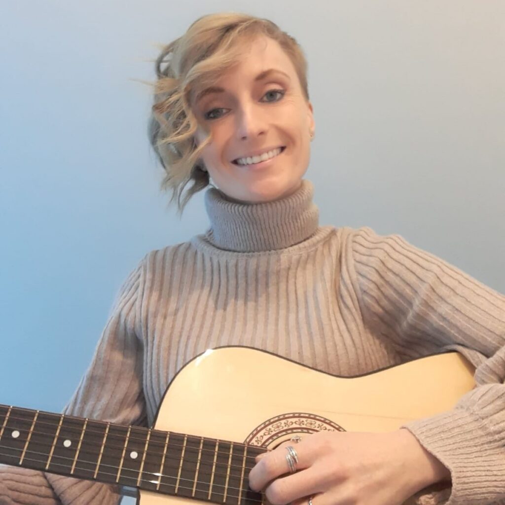 Karen: Picture shows a women with short blonde wavey hair smiling. She is wearing a beige coloured turtleneck top and playing an acoustic guitar