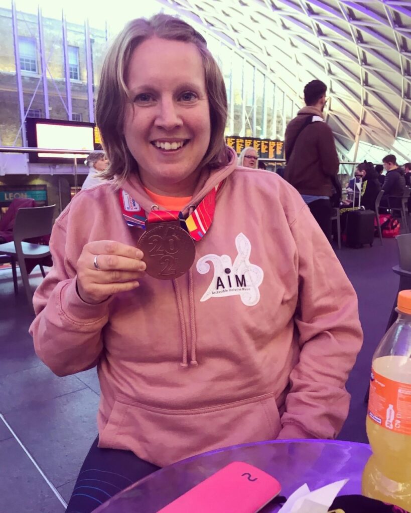 Sarah: Picture shows a women with mid-length fair hair smiling and holding a running medal. She is wearing a pink hoodie with the Accessible Inclusive Music logo