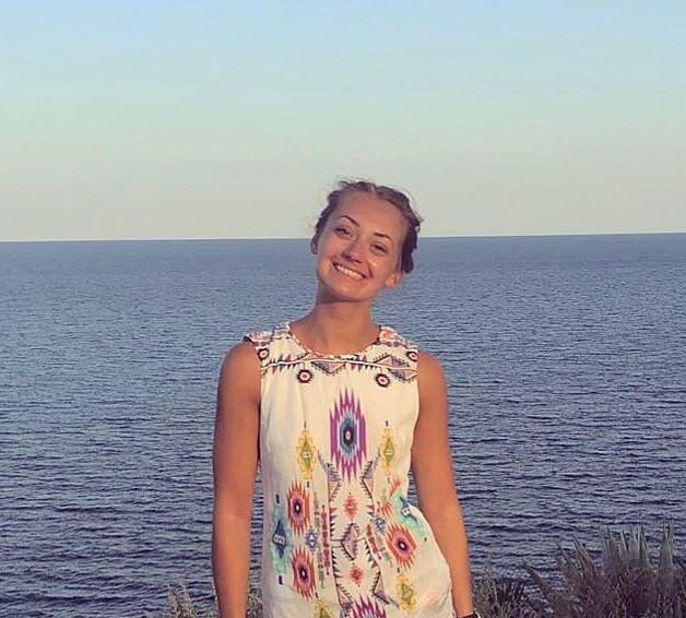 Chloe: picture shows a women standing in front of the sea smiling. She has brown hair which is tied back and a white dress with colourful shapes on it.