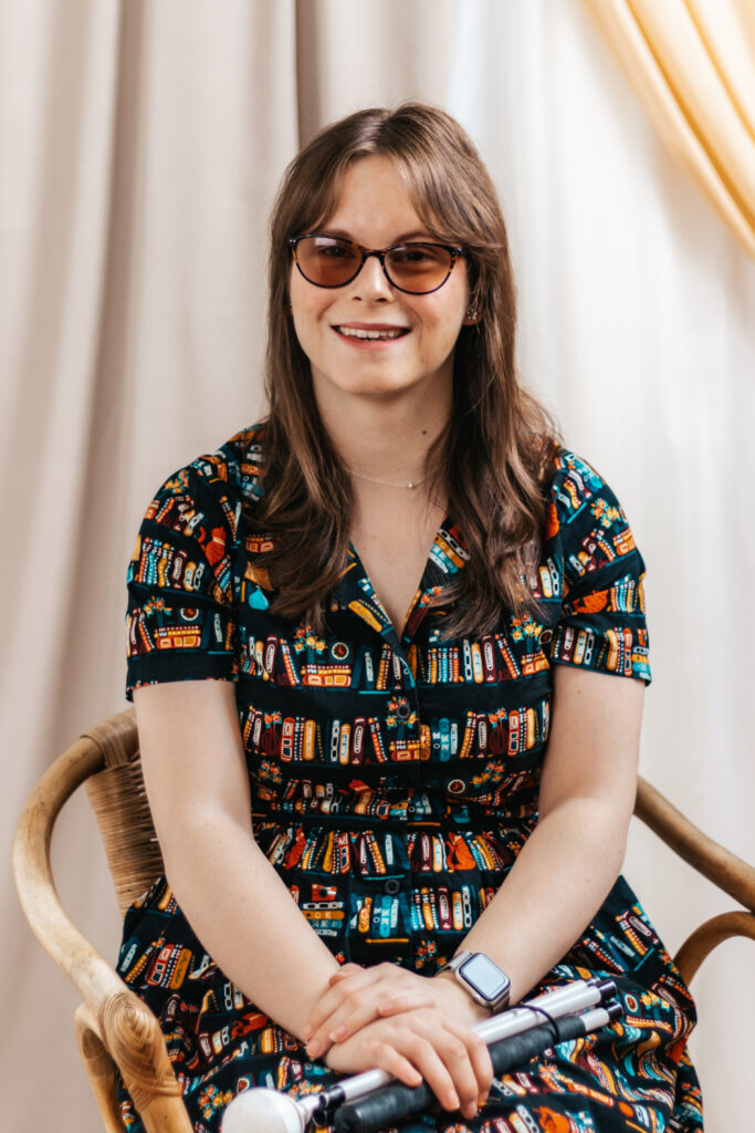 Chloe: Picture shows a women with long brown hair and glasses smiling. She is wearing a black dress with a repeated multicolour book pattern. She is holding a white cane which is folded on her lap.