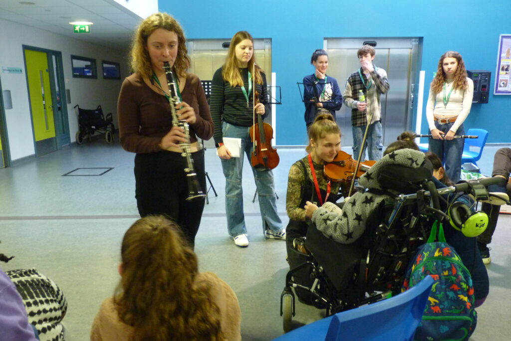 A range of students playing different instruments. One person is in a wheelchair waiting the students. 