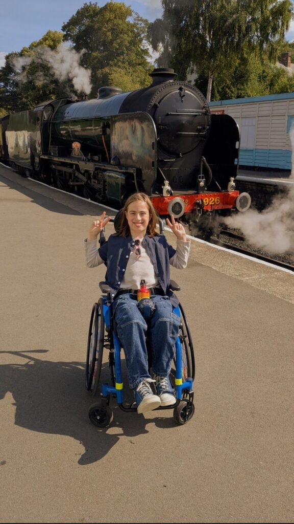 A boy sat in a blue wheelchair. He’s wearing jeans and a top and is looking at the camera. 